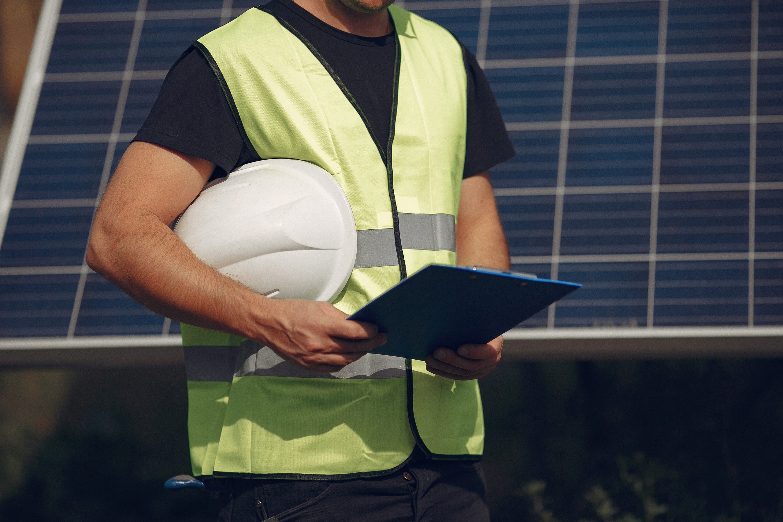 Um trabalhador com vestuário de segurança está com um capacete e uma prancheta, diante de um painel solar, sugerindo que ele está realizando tarefas relacionadas à energia solar. O rosto do trabalhador não é visível.