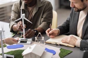 uas pessoas estão examinando uma maquete de energia renovável, com uma focando em um modelo de turbina eólica e a outra anotando em um caderno.