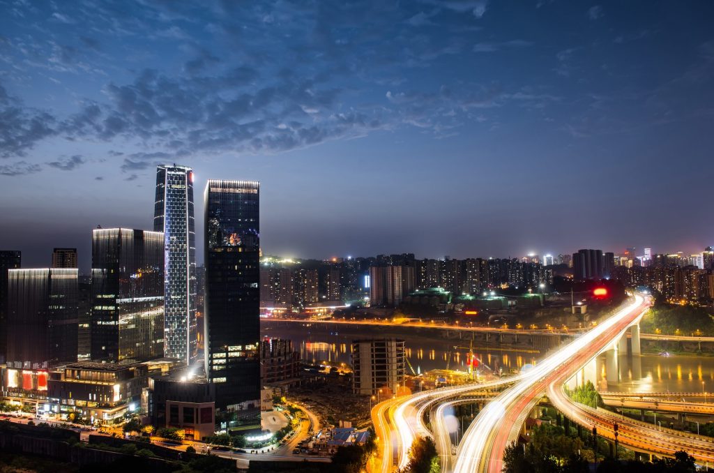 Vista noturna de uma cidade moderna com arranha-céus iluminados e um complexo de viadutos com luzes de tráfego brilhantes, destacando a energia vibrante da vida urbana.