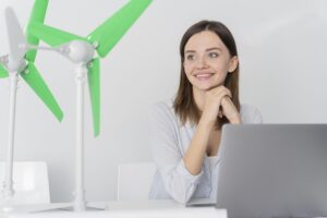 Mulher sorrindo em um escritório ao lado de um modelo de turbina eólica verde, representando energia renovável e inovação sustentável.