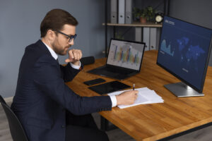 Imagem de um assessor analisando um contrato de energia para renegociar com as melhores condições. Na imagem vemos um homem de terno sentado atrás de uma mesa de madeira escrevendo em um papel com o computador ligado.